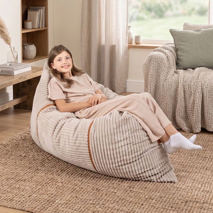 A young girl wearing beige pajamas sits comfortably on a large ribbed beige bean bag chair in a cozy, sunlit living room. 