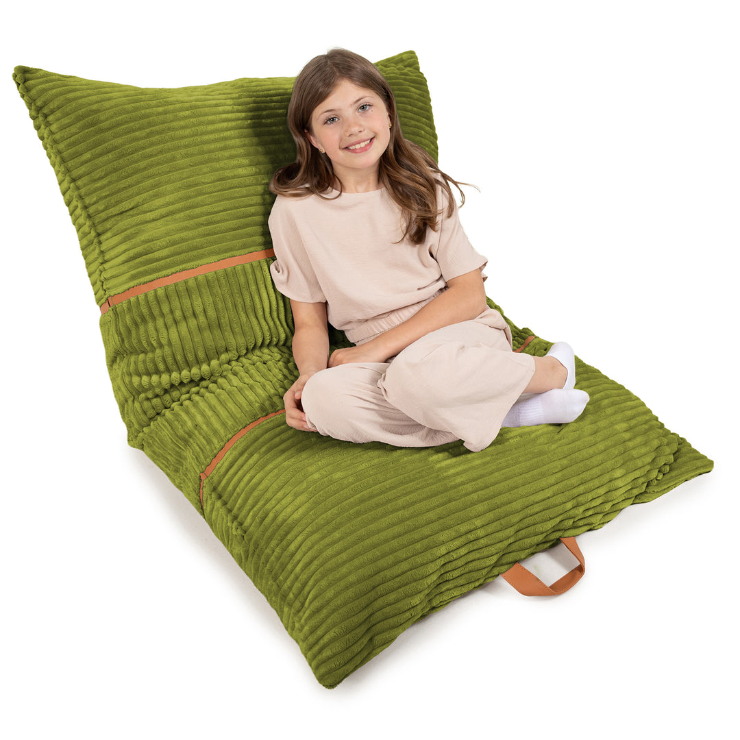 A young girl with long brown hair sits cross-legged on a large, olive green corduroy floor pillow. The pillow is angled diagonally and features a brown leather strap handle on the bottom right. 