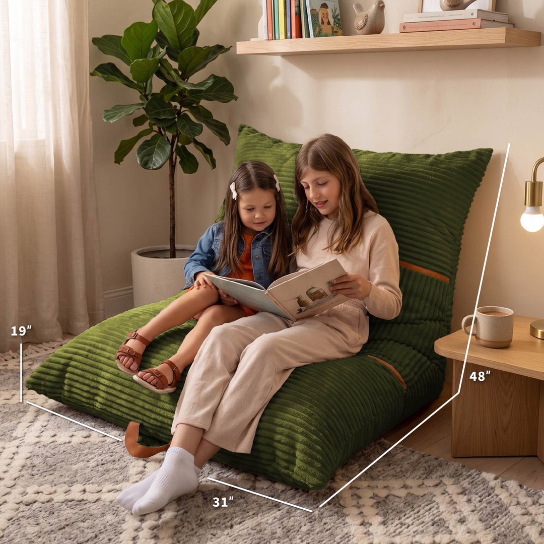 A woman and a child sit on a large green ribbed floor cushion in a cozy living room setting, reading a book. White dimension lines overlay the cushion showing measurements of 19 inches, 31 inches, 48 inches, and 35 inches. 
