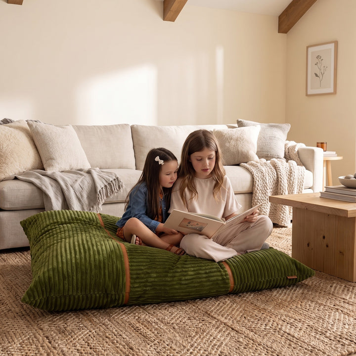 Two children sitting on a large olive green ribbed floor cushion in a cozy, sunlit living room, reading a book. The room features a cream-colored sofa, a wooden coffee table, and a textured jute rug. 