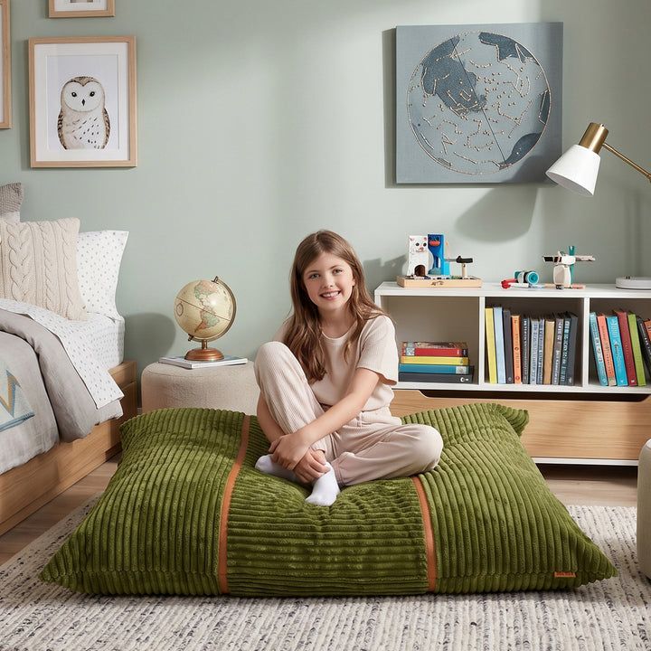 A young girl sits cross-legged on a large olive green corduroy floor cushion in a styled bedroom setting. The room features a light sage green wall, a wooden bed, a bookshelf, and a globe on a side table. 