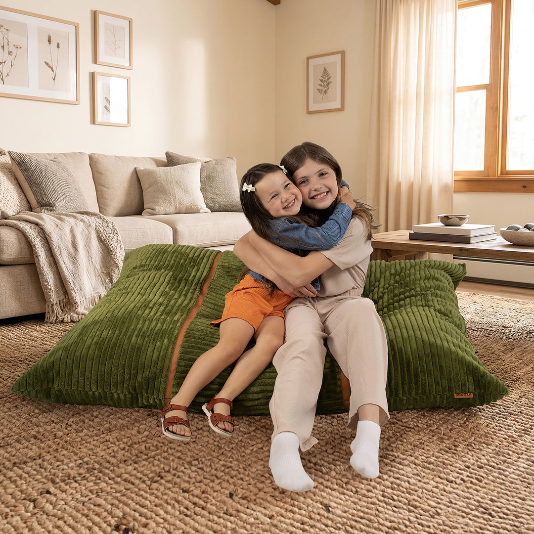 Two children hugging while sitting on a large green ribbed floor cushion in a warm, modern living room with a beige sofa and woven rug. 
