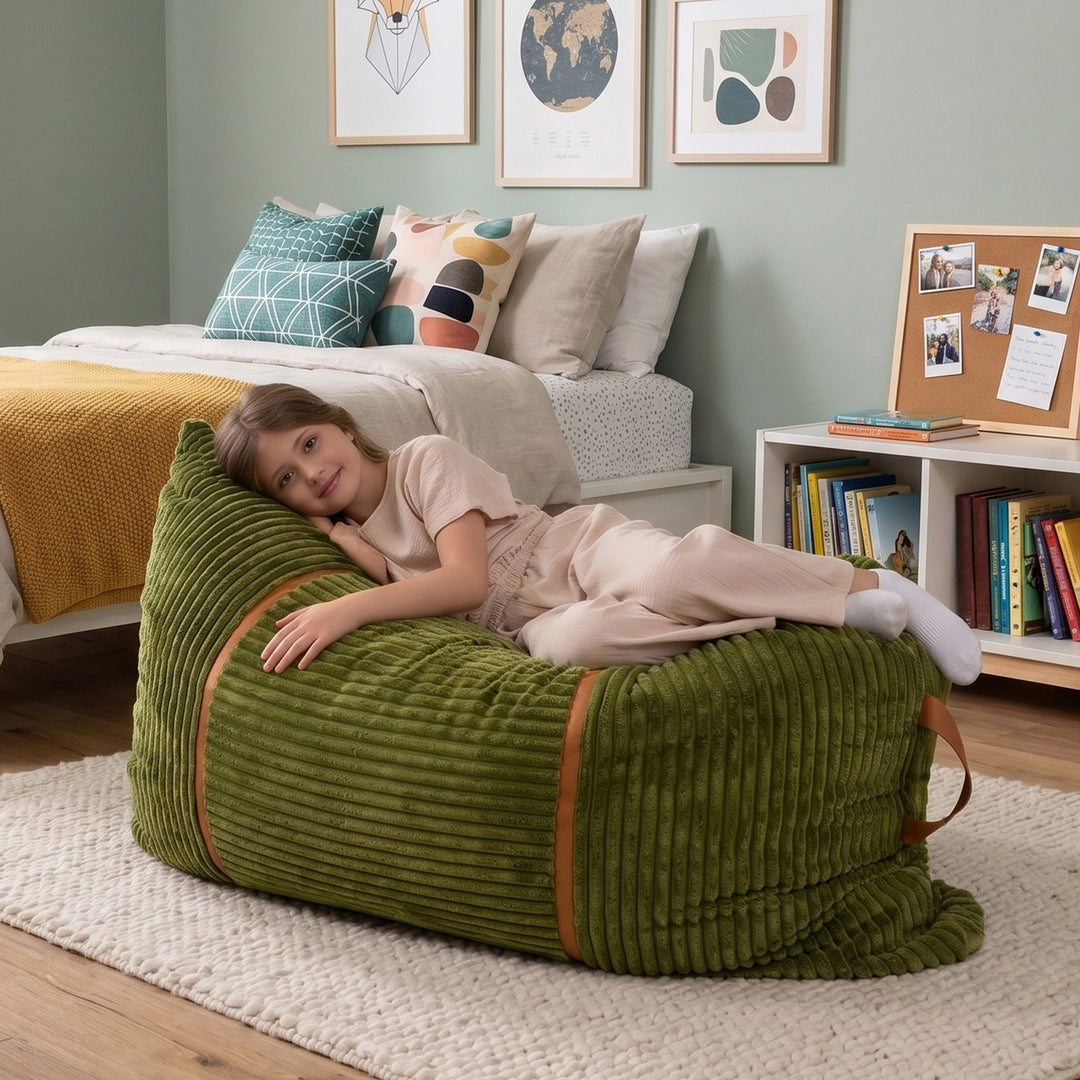 A child lies on a large olive green corduroy bean bag chair in a styled bedroom setting. The bean bag has brown leather-like trim and is placed on a white rug in front of a bed with neutral-toned pillows and framed wall art. #color_moss-green-corded-plush