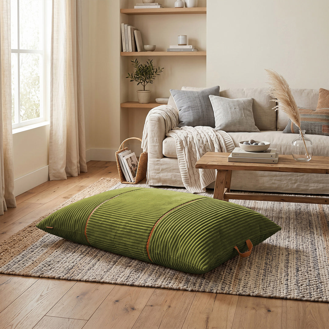 An olive green ribbed corduroy floor pillow with brown leather handles, resting on a woven area rug in a bright, modern living room. 