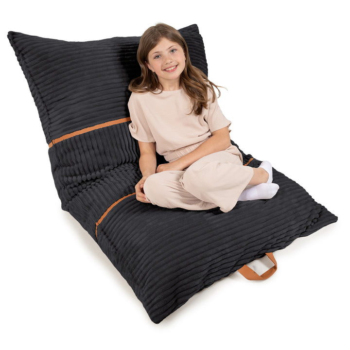 A young girl sits cross-legged on a large black ribbed floor pillow with tan accents, photographed on a clean white background. 
