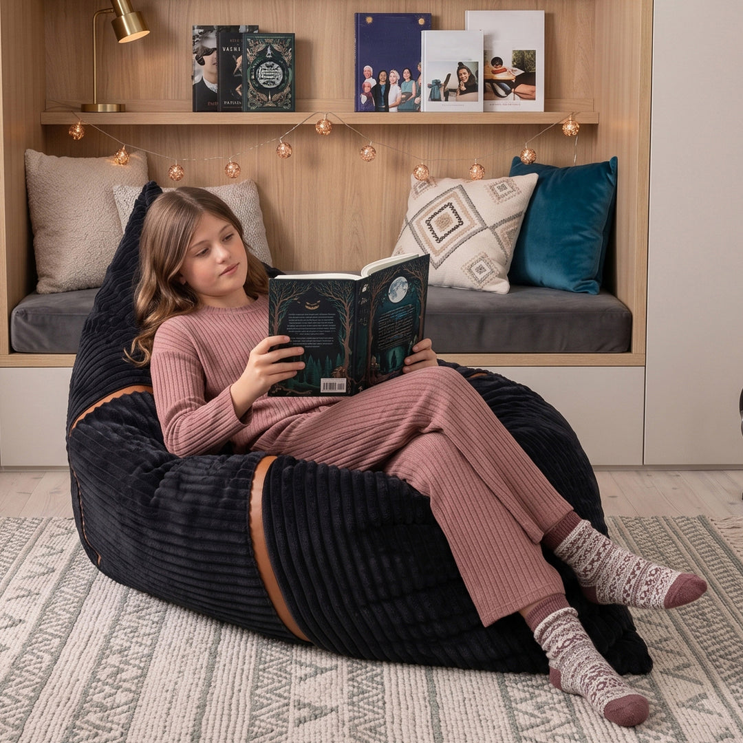 A young girl in a matching pink ribbed pajama set relaxes in a large black corduroy bean bag chair, reading a book in a cozy bedroom setting. 