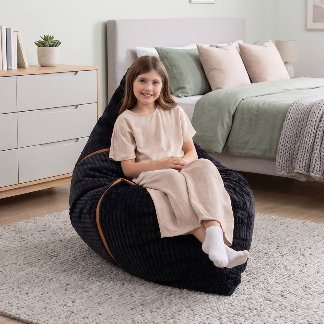 A young girl with brown hair sits smiling in a black ribbed corduroy bean bag chair with brown trim, placed on a light gray rug in a modern bedroom setting. 