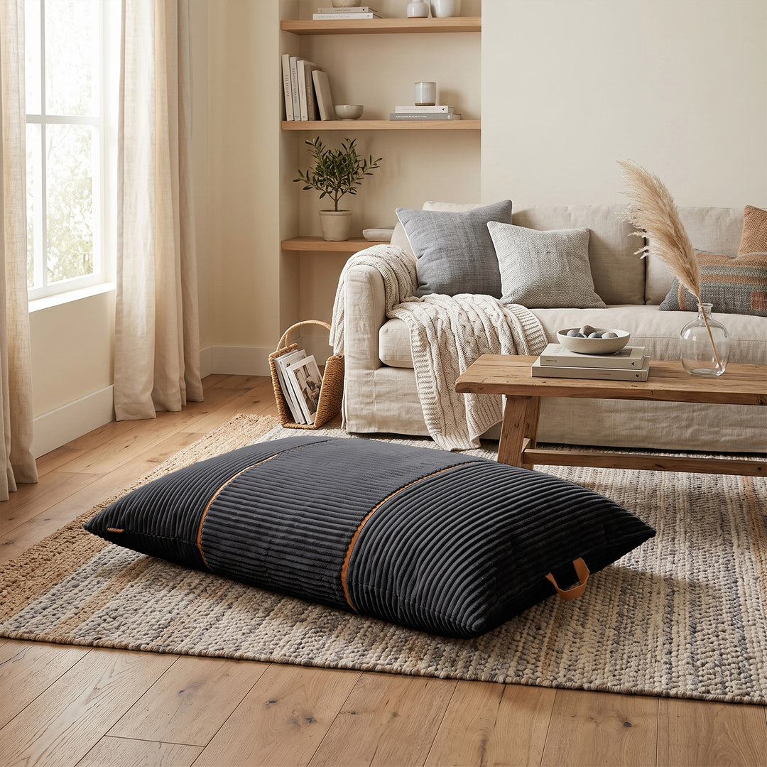 A dark charcoal gray ribbed floor pillow with orange-brown piping details, resting on a woven jute rug in a modern, warm-toned living room setting. 