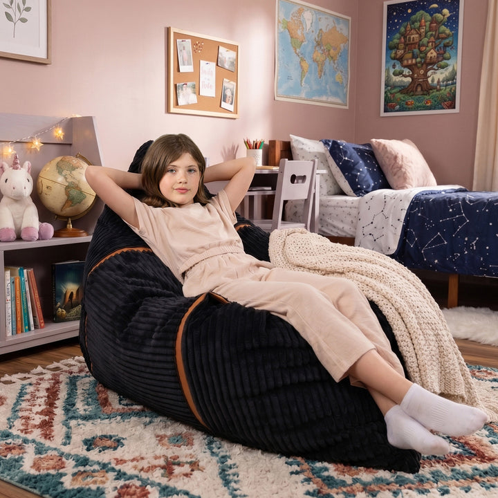 A child wearing a beige pajama set reclines on a large black ribbed bean bag chair in a decorated bedroom. The bean bag has thin brown trim and is placed on a patterned rug, with a bed, books, and framed wall art visible in the background. 