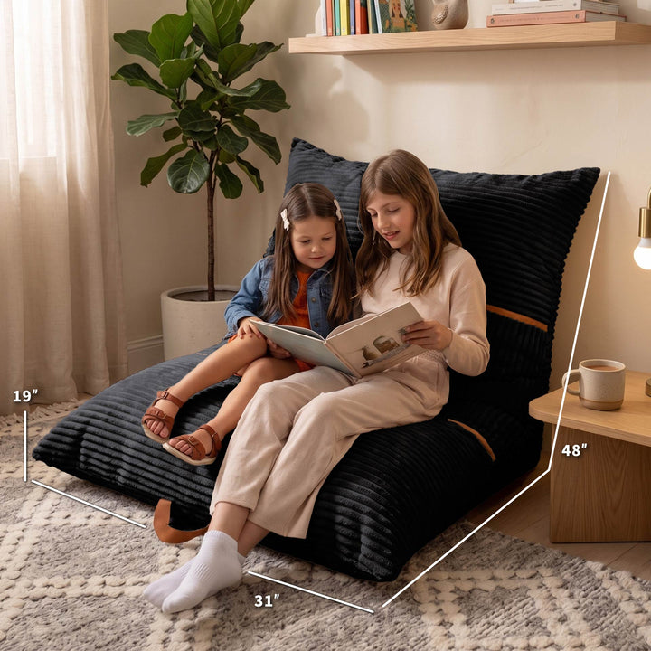 Two girls sitting on a large black ribbed floor cushion in a cozy room, reading a book. White dimension lines overlay the cushion showing measurements of 19 inches, 31 inches, 48 inches, and 35 inches. 