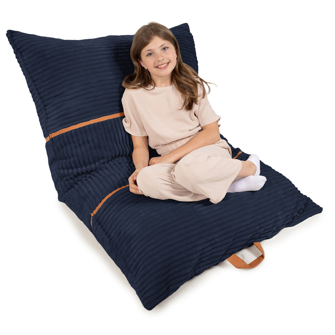 A young girl with long brown hair sits cross-legged on a large, ribbed navy blue floor cushion with tan accents, set against a solid white background. 