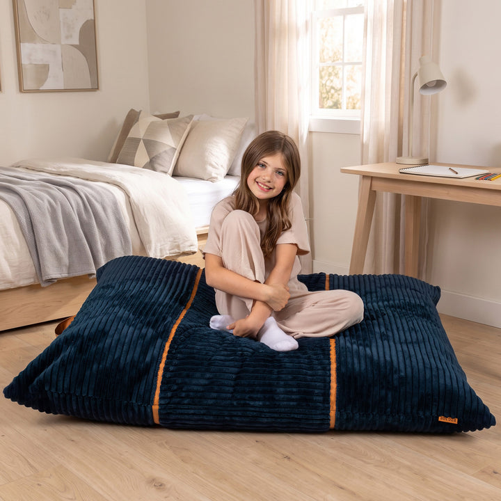 A young girl with long brown hair sits smiling on a large, deep navy blue corduroy floor cushion with orange piping details, set in a bright, modern bedroom. 