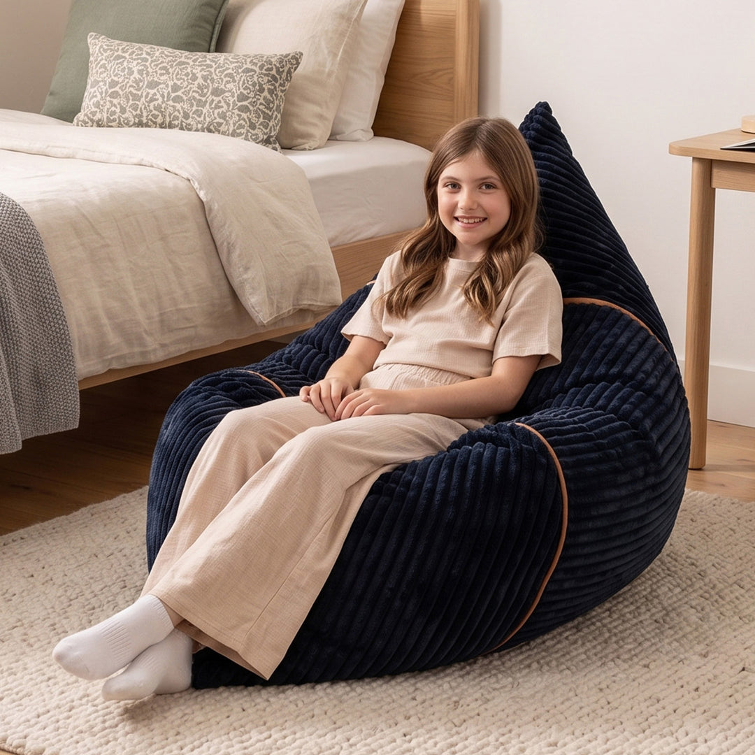 A young girl with long brown hair sits smiling in a dark navy blue corduroy bean bag chair with tan piping, placed on a light-colored rug in a bedroom setting. 