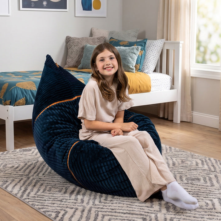A young girl with long brown hair sits smiling on a large, plush navy blue bean bag chair in a bright, modern bedroom. The bean bag features subtle orange piping accents. 