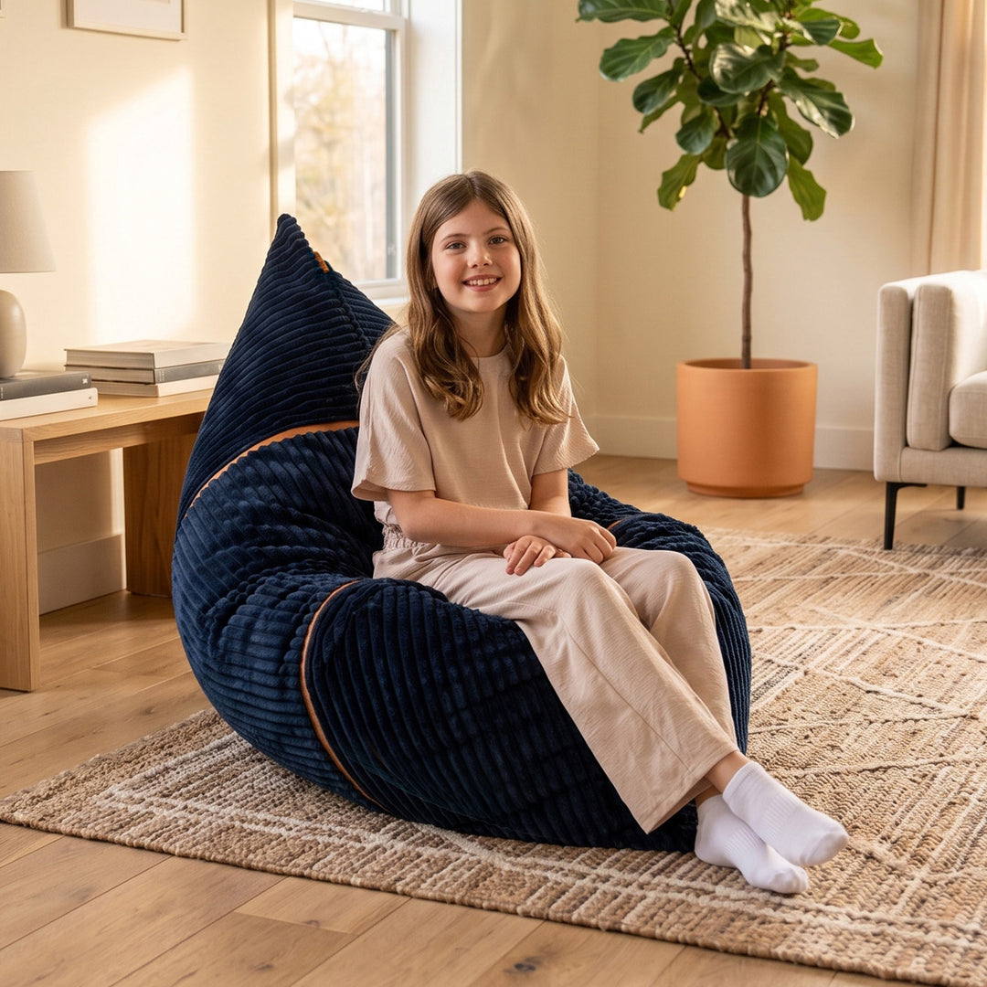 A young girl with long light brown hair sits smiling on a large, plush navy blue bean bag chair in a cozy, sunlit living room. The bean bag has subtle tan piping details. 
