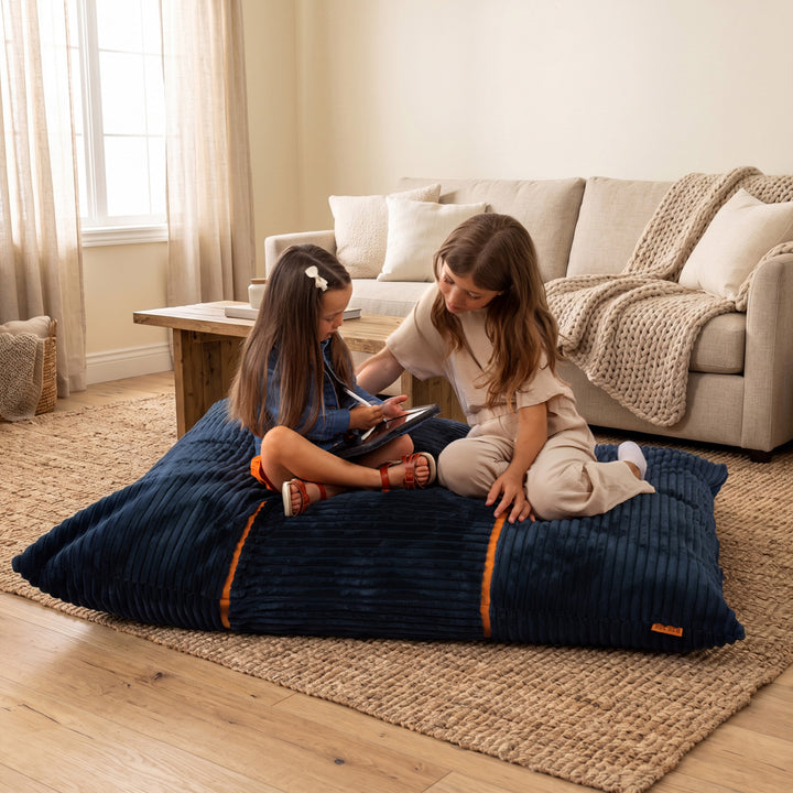 Two children sitting on a large navy blue corduroy floor cushion in a cozy living room setting with a beige sofa and wooden floor. 