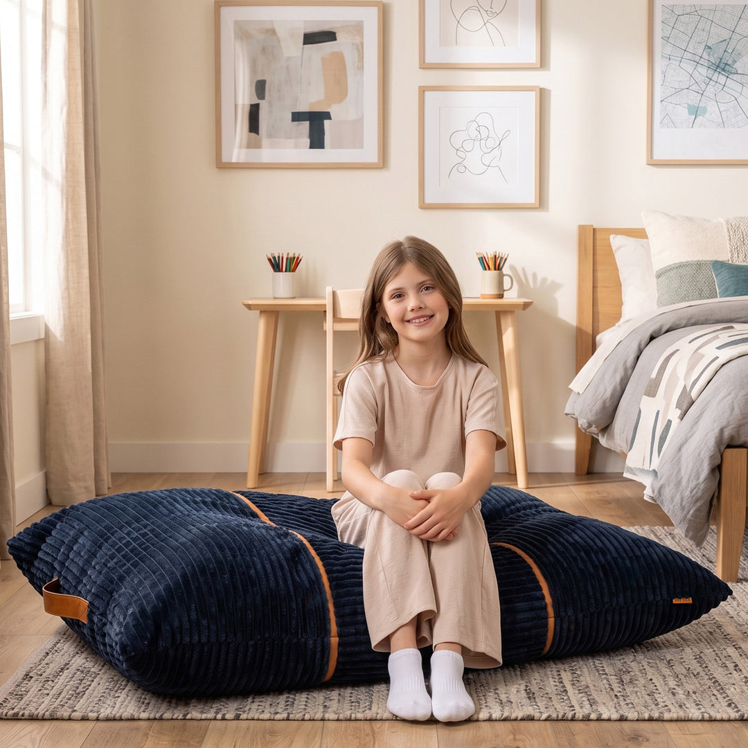 A young girl with long brown hair sits smiling on a large navy blue corduroy floor cushion in a cozy, modern bedroom. The cushion features thin orange-brown piping accents. 
