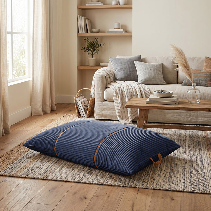 A large rectangular navy blue ribbed floor cushion with tan leather-like corner accents, placed on a woven area rug in a styled living room with a sofa, coffee table, and wood floor. 