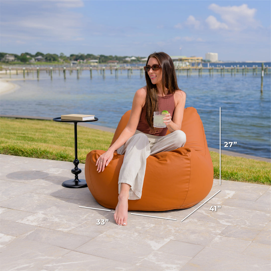 Woman sitting on a bean bag chair by a waterfront with measurements displayed. #color_honey-sand-outdoor-vegan-leather