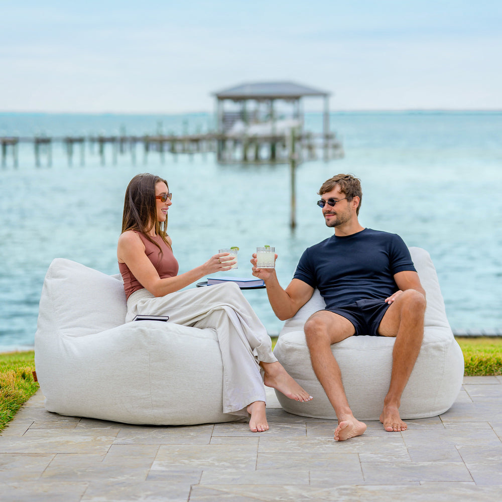 Two people sitting on bean bags by the water, holding drinks. #color_sunlit-ivory-outdoor-performance-linen