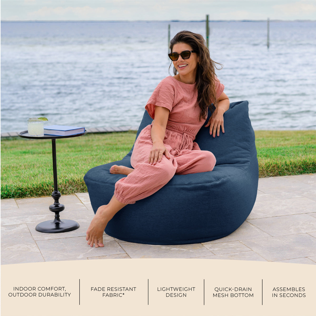 Woman sitting on a navy bean bag chair by a waterfront, with text about product features. #color_sailor-blue-outdoor-performance-linen