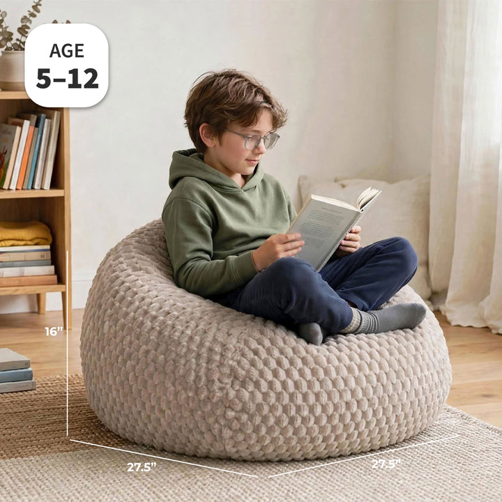 A child sits cross-legged on a large, round, chunky bean bag chair in a cozy room setting, reading a book. White dimension lines and text overlays indicate the chair&