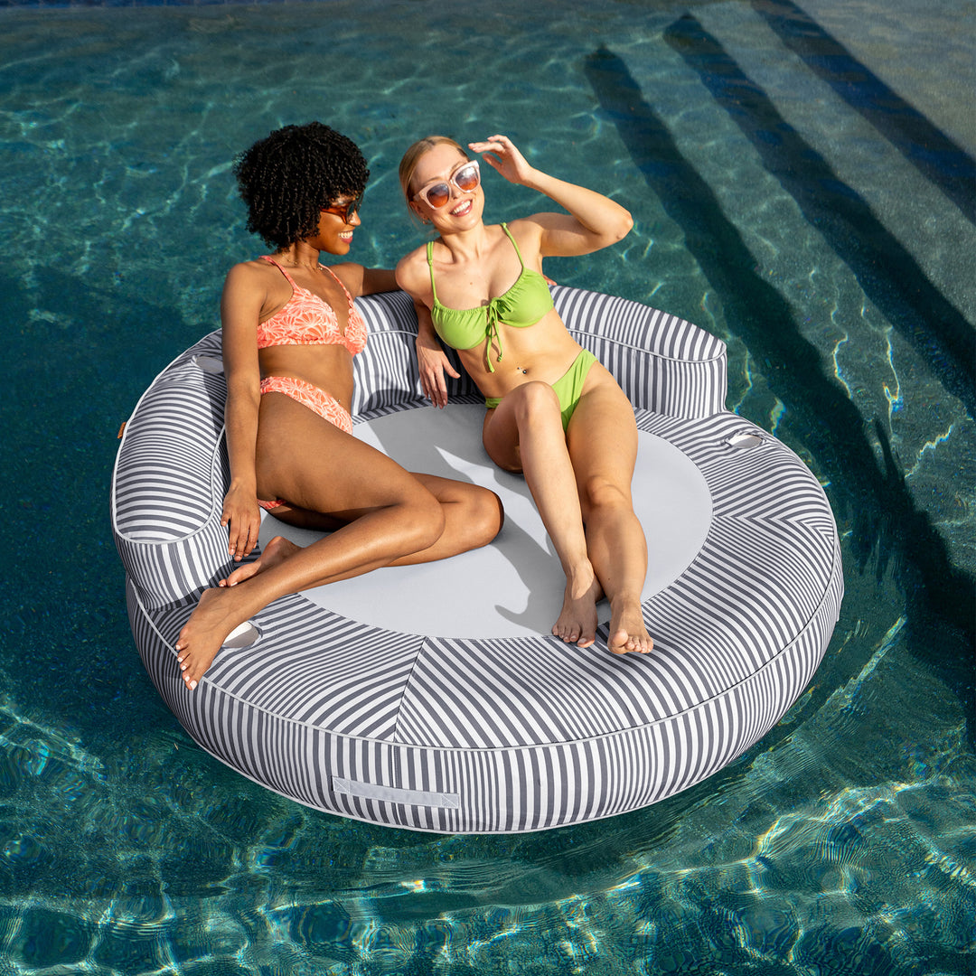 Two women in swimsuits sitting on a circular pool float in a pool. #color_shoreline-gray-stripe-olefin