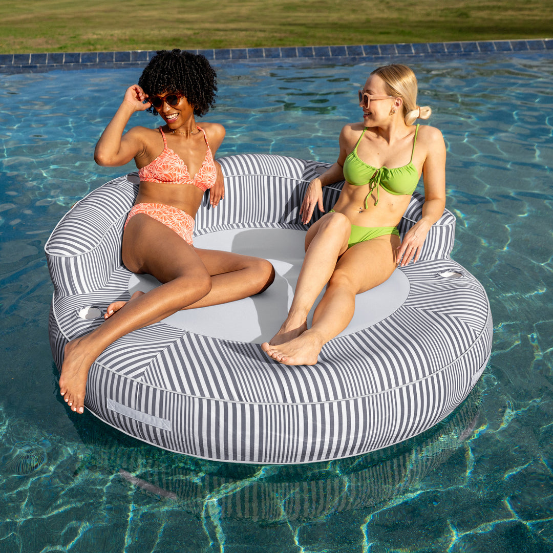 Two women sitting on a striped pool float in a pool #color_shoreline-gray-stripe-olefin