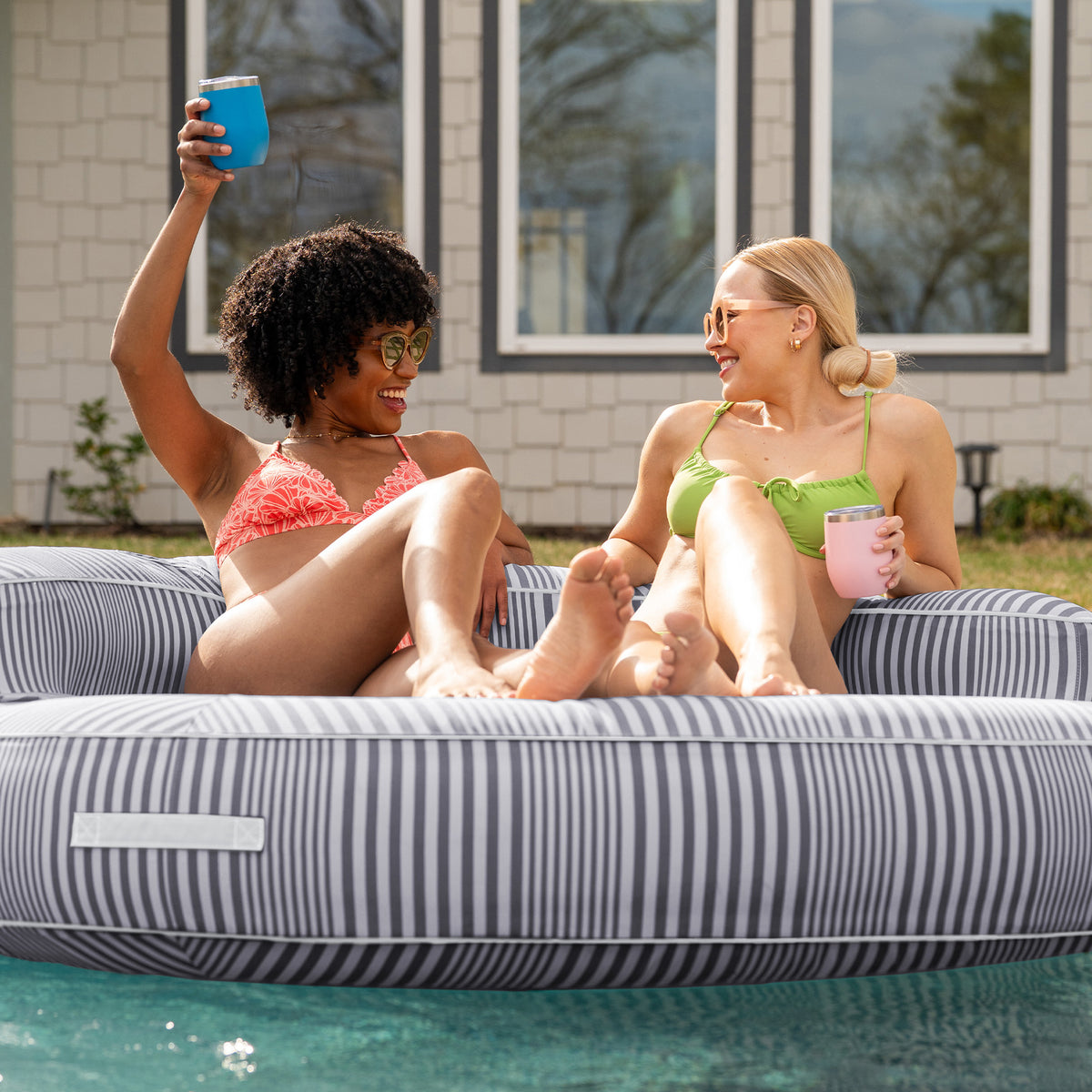 Two women sitting on a striped pool float in a pool, enjoying drinks. 