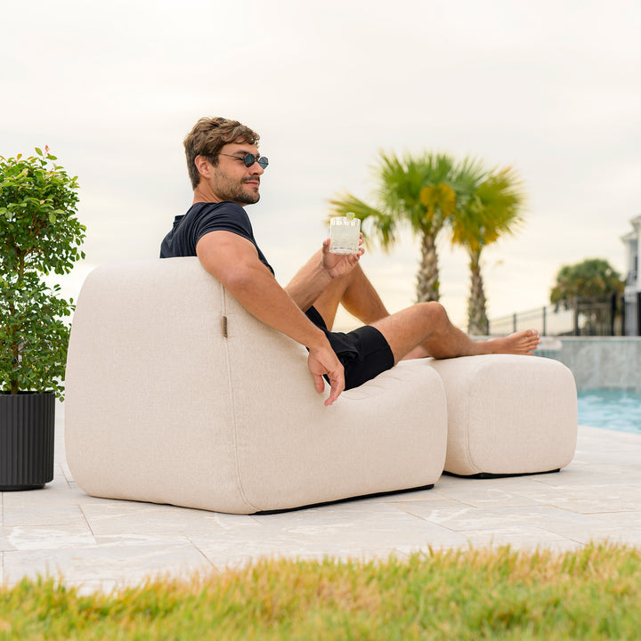 Man sitting on a beige outdoor lounge chair by a pool with palm trees in the background #color_sunlit-cream-outdoor-performance-linen