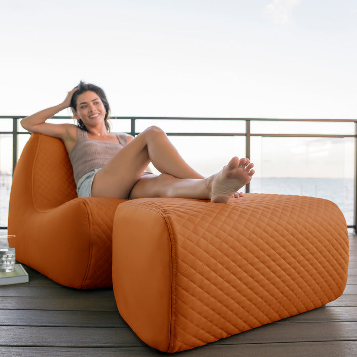 Woman relaxing on a large orange chair by a body of water. #color_honey-sand-outdoor-vegan-leather
