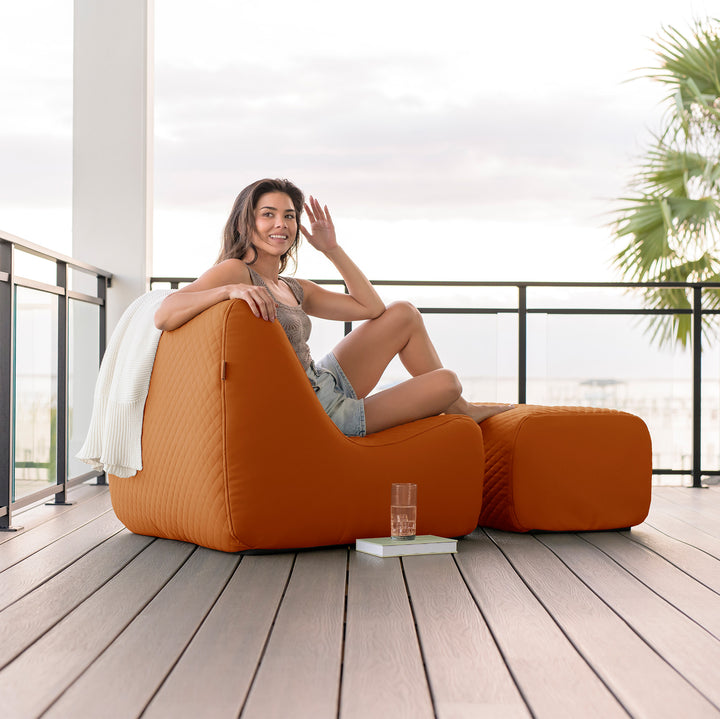 Woman sitting on a large orange chair on a wooden deck with a white wall and plant in the background. #color_honey-sand-outdoor-vegan-leather