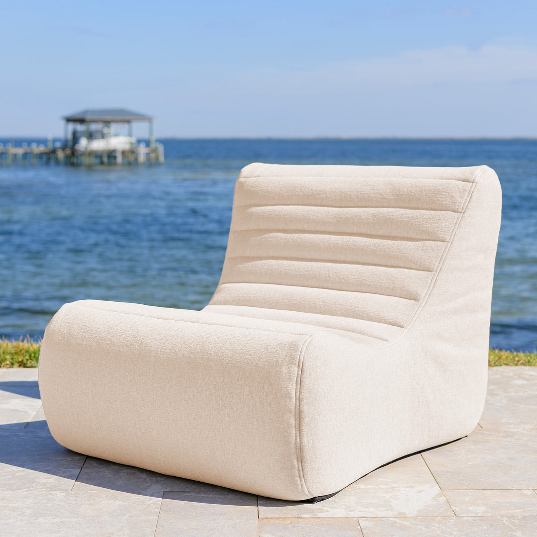 Beige lounge chair by the sea with a clear blue sky. #color_sunlit-cream-outdoor-performance-linen