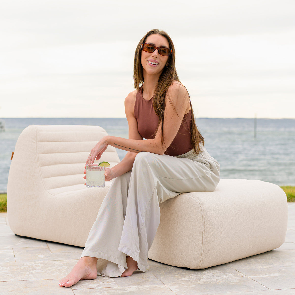 Woman sitting on a modern outdoor chair by the water, holding a drink. 