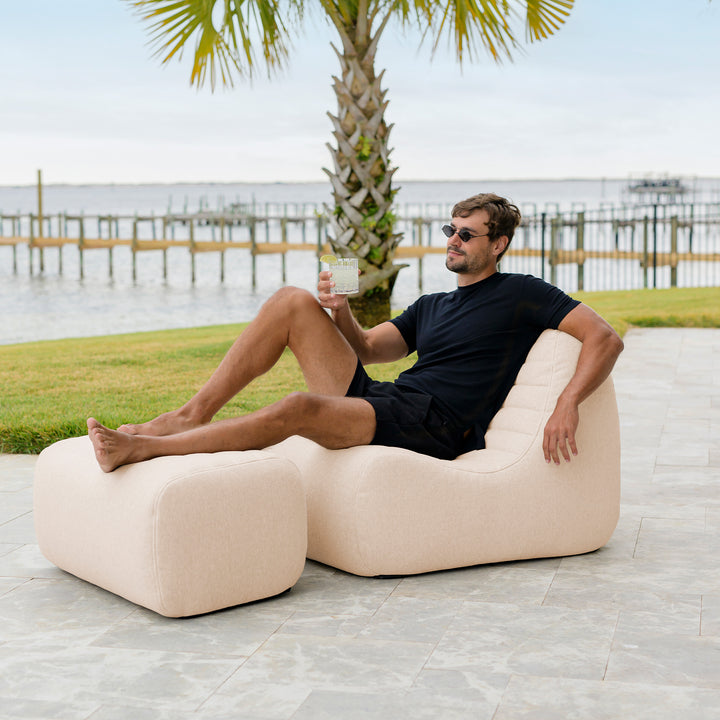 Man relaxing on a beige outdoor sofa by a waterfront with palm trees in the background #color_sunlit-cream-outdoor-performance-linen