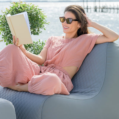 Woman in a pink dress reading a book on a gray sofa by a waterfront. 