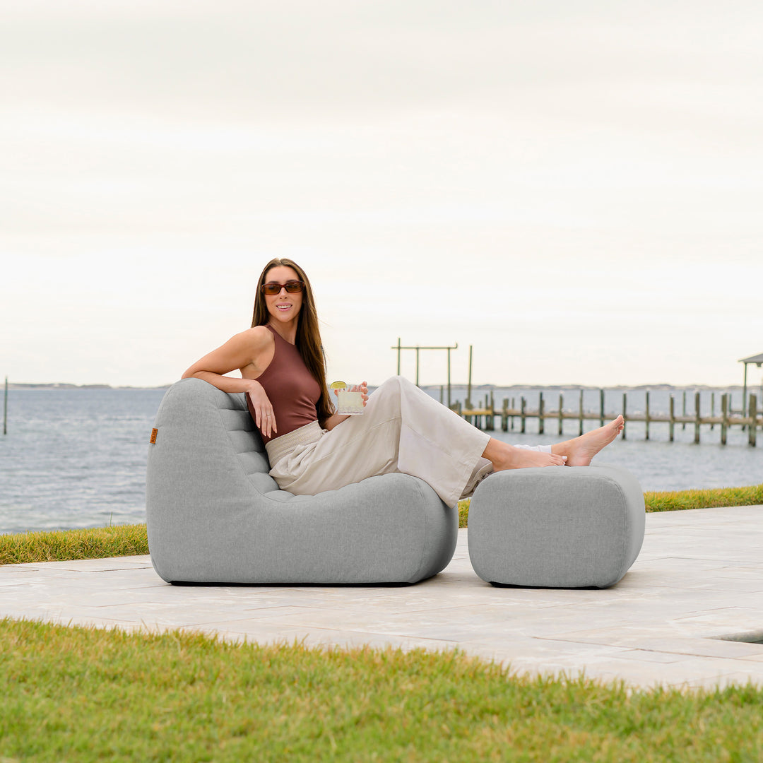 Woman sitting on a gray outdoor chair by a waterfront. #color_port-gray-outdoor-performance-linen