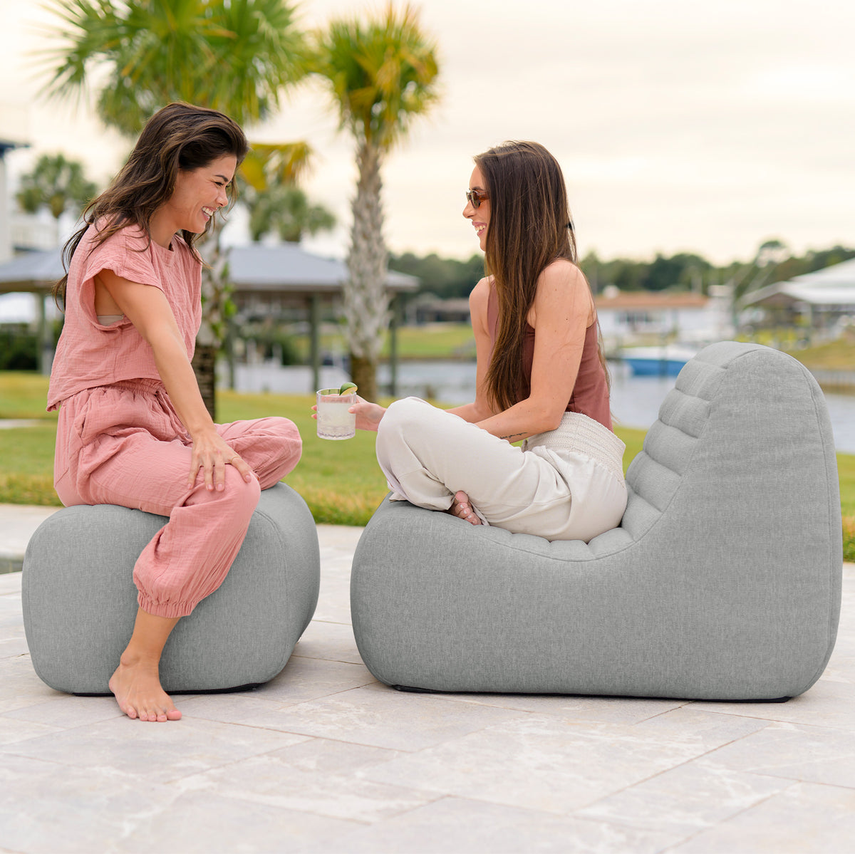 Two women sitting on gray chairs outdoors with palm trees and a lake in the background. 