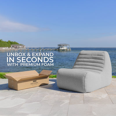 Gray outdoor chair next to a cardboard box with text 'Unbox & Expand in Seconds with Premium Foam' by Solvana, set against a scenic waterfront background. 