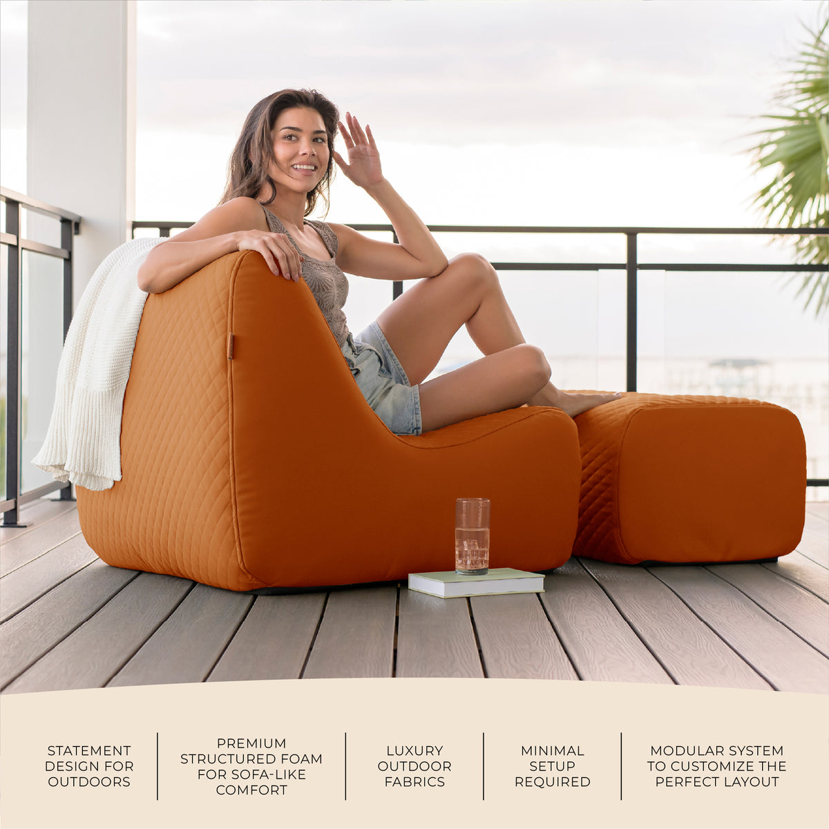 Woman sitting on a large orange patio chair on a wooden deck with a view of the ocean. 