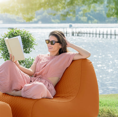 Woman in a pink dress reading a book on an orange chair by a lake. 