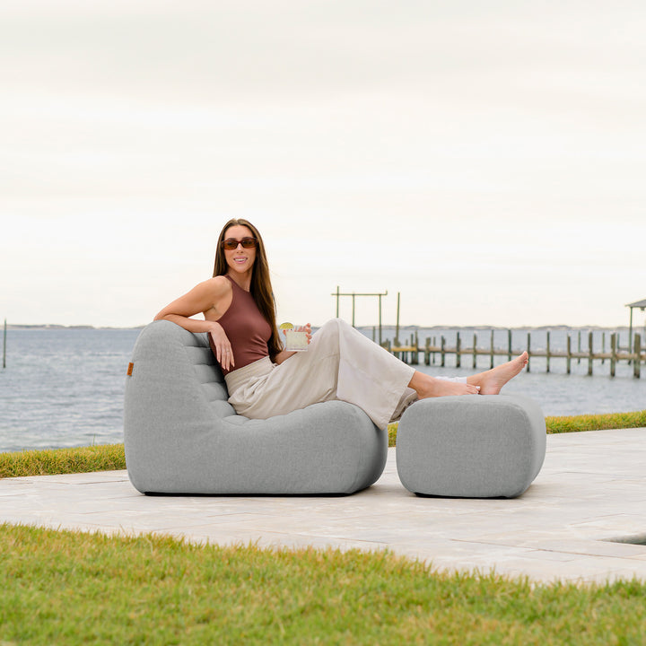 Woman sitting on a gray outdoor chair by a waterfront. #color_port-gray-outdoor-performance-linen