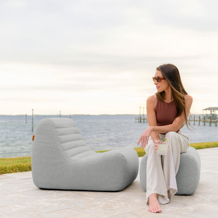 Woman sitting on a modern outdoor chair by the water #color_port-gray-outdoor-performance-linen