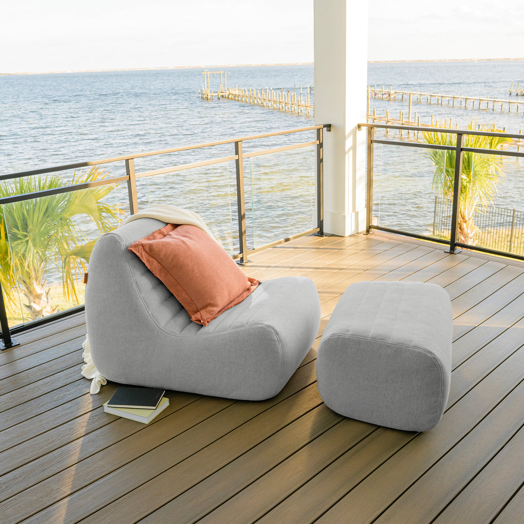 Gray lounge chair with a pink pillow on a wooden deck by the water #color_port-gray-outdoor-performance-linen