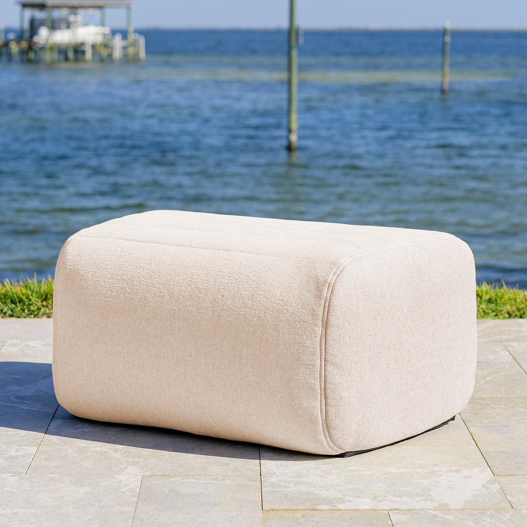 Beige ottoman on a patio with water and dock in the background #color_sunlit-cream-outdoor-performance-linen