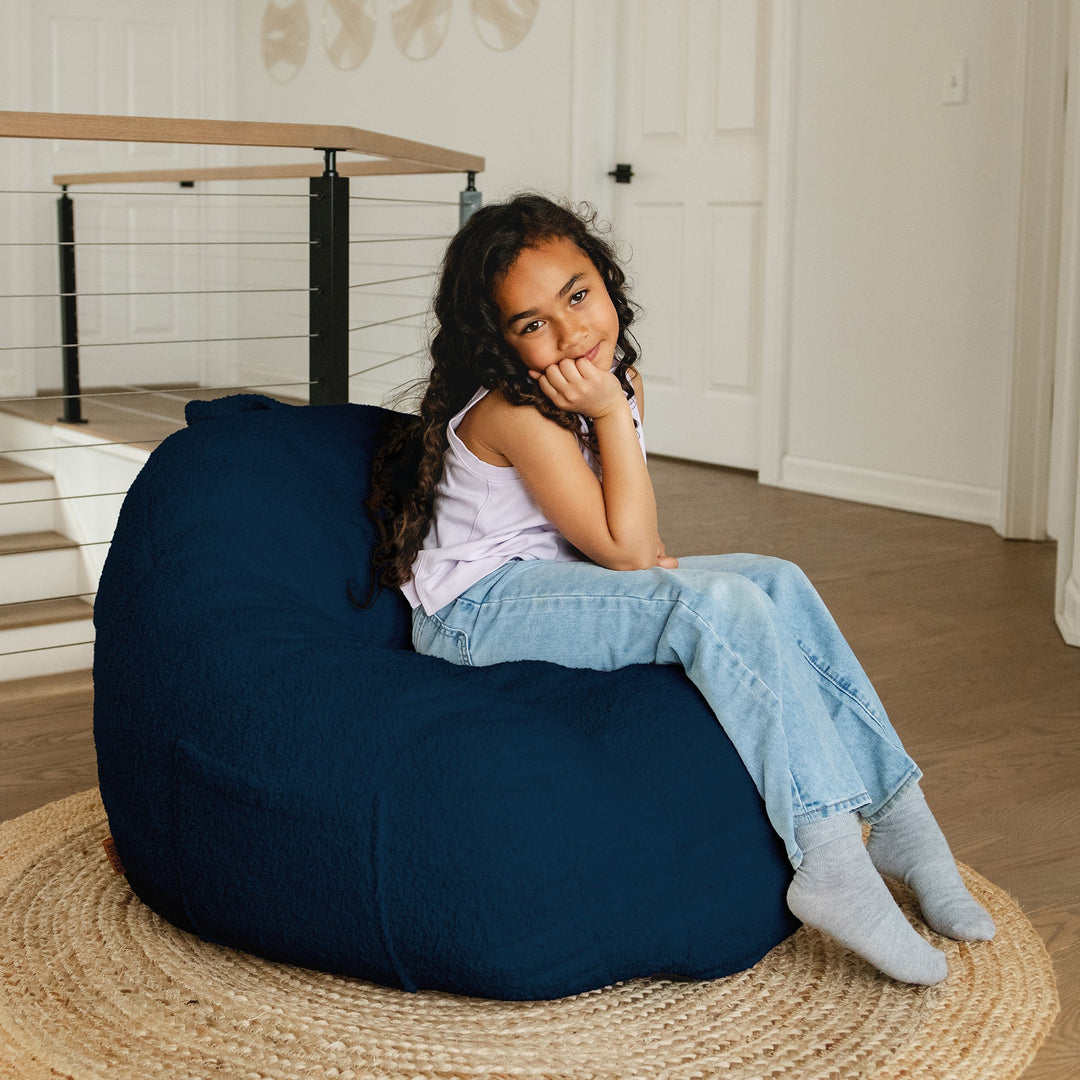Young girl sitting on a navy bean bag chair in a home setting. #color_deep-blue-teddy-boucle
