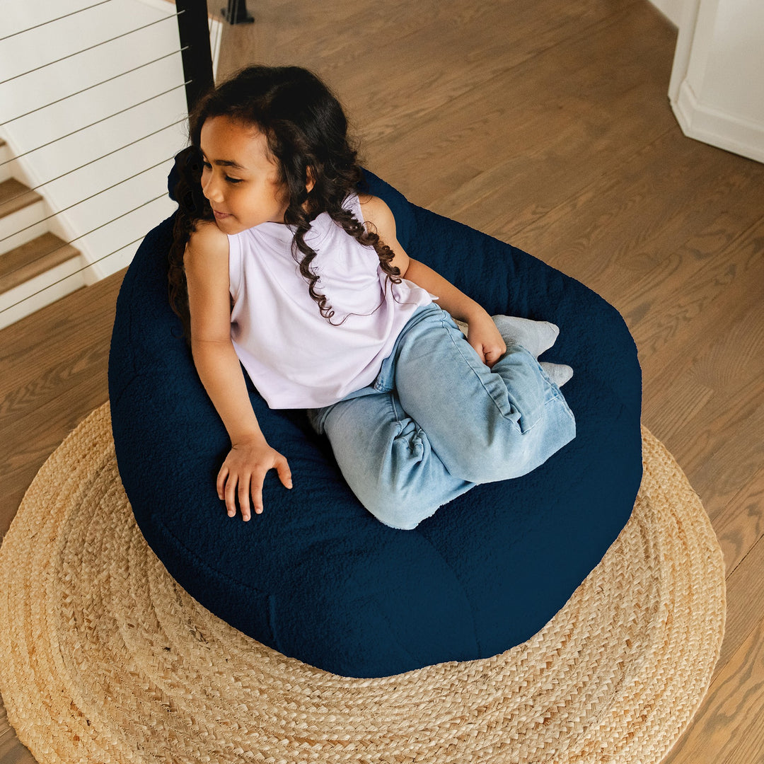 Child sitting on a navy bean bag chair in a home setting #color_deep-blue-teddy-boucle