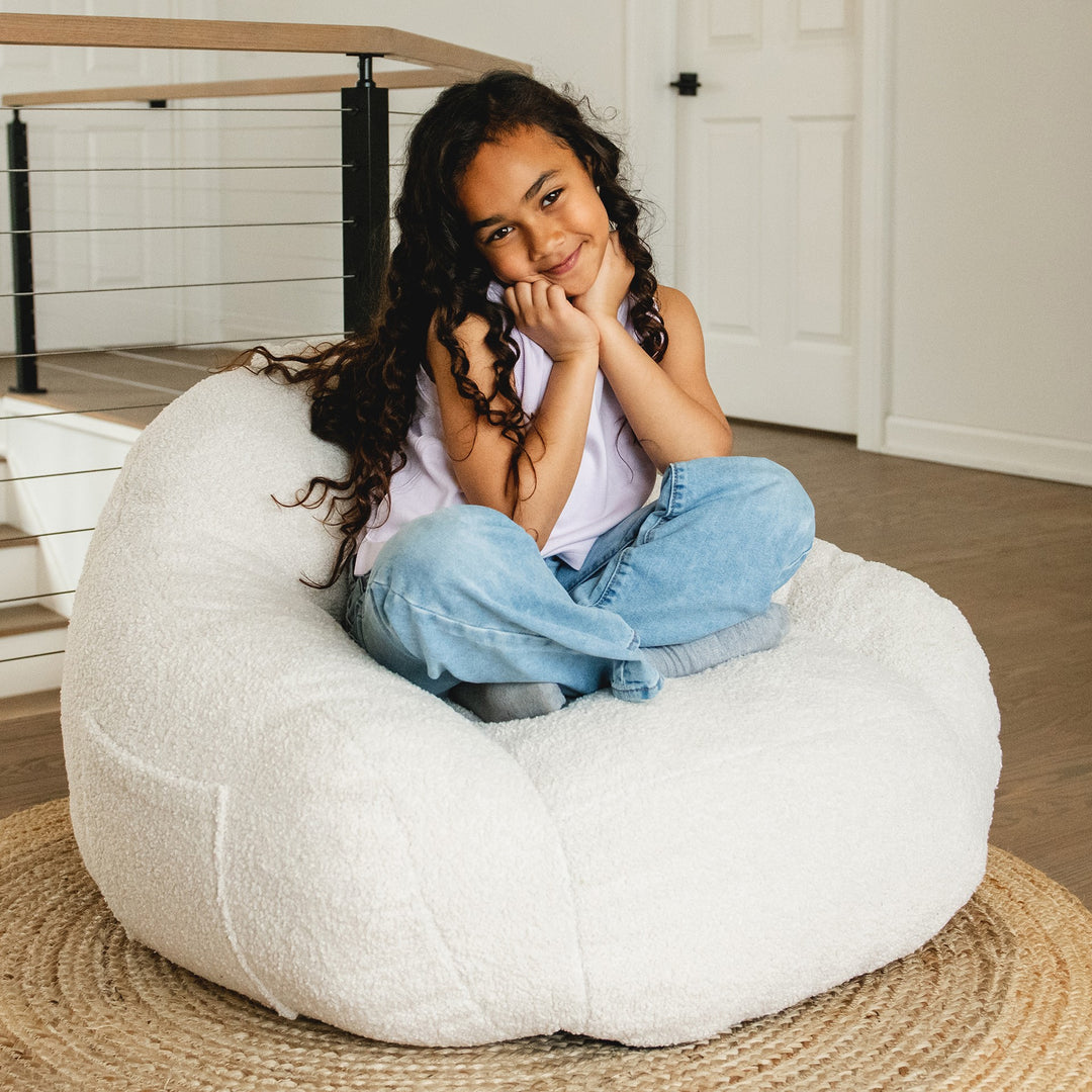 Young girl sitting on a white bean bag chair in a room with wooden floor and white walls. #color_icing-teddy-boucle