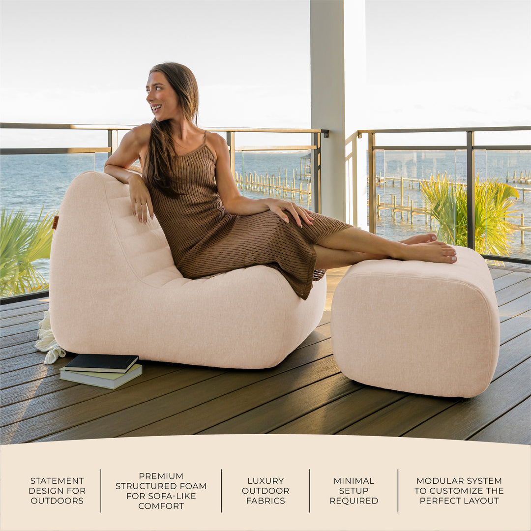 Woman sitting on a beige chair with a matching ottoman on a deck with ocean view. #color_sunlit-cream-outdoor-performance-linen