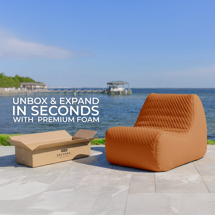 Brown outdoor chair with a cardboard box on a patio by a waterfront, with text about quick assembly. #color_honey-sand-outdoor-vegan-leather